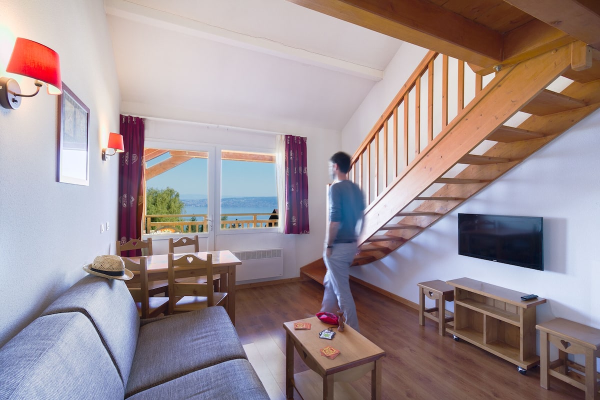 A living room with a wooden staircase, a TV mounted on the wall, and a view of the outdoors through large windows.