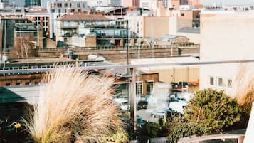 A rooftop terrace with wooden furniture, a table with a plate of food and drinks, and a view of a cityscape.