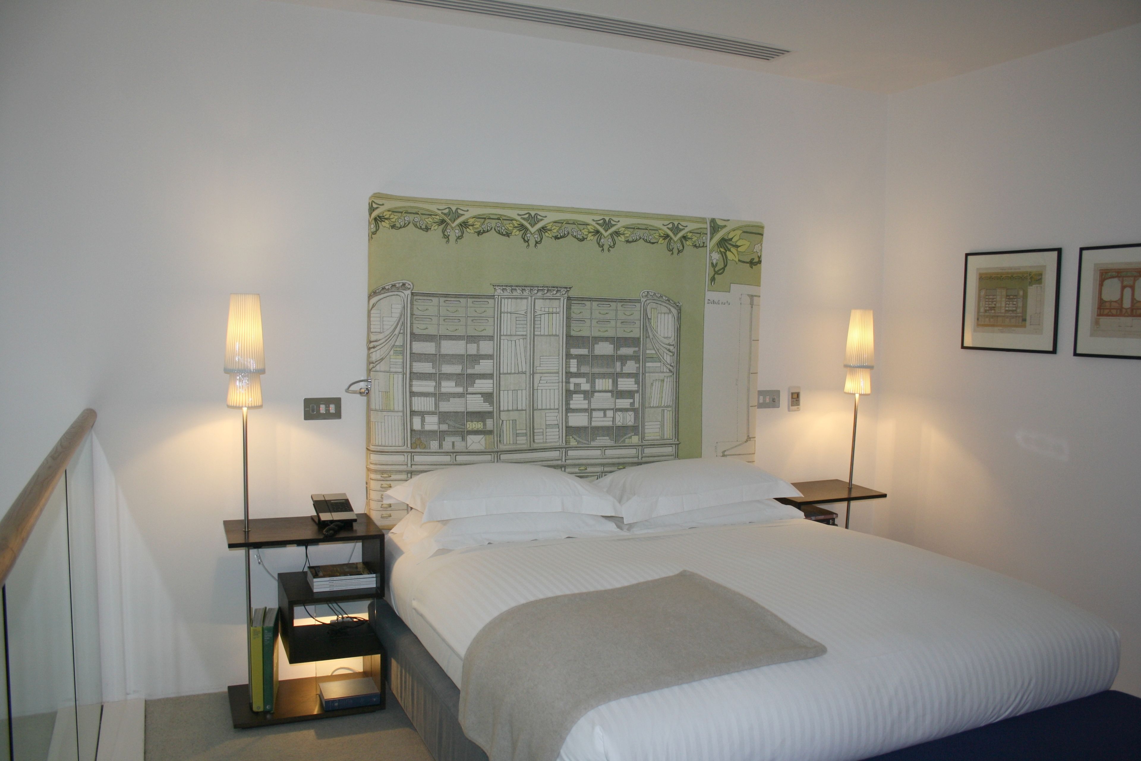 Een net opgemaakt bed met witte lakens, twee nachtkastjes met lampen, een boekenkast en een muur met kunstwerken.
