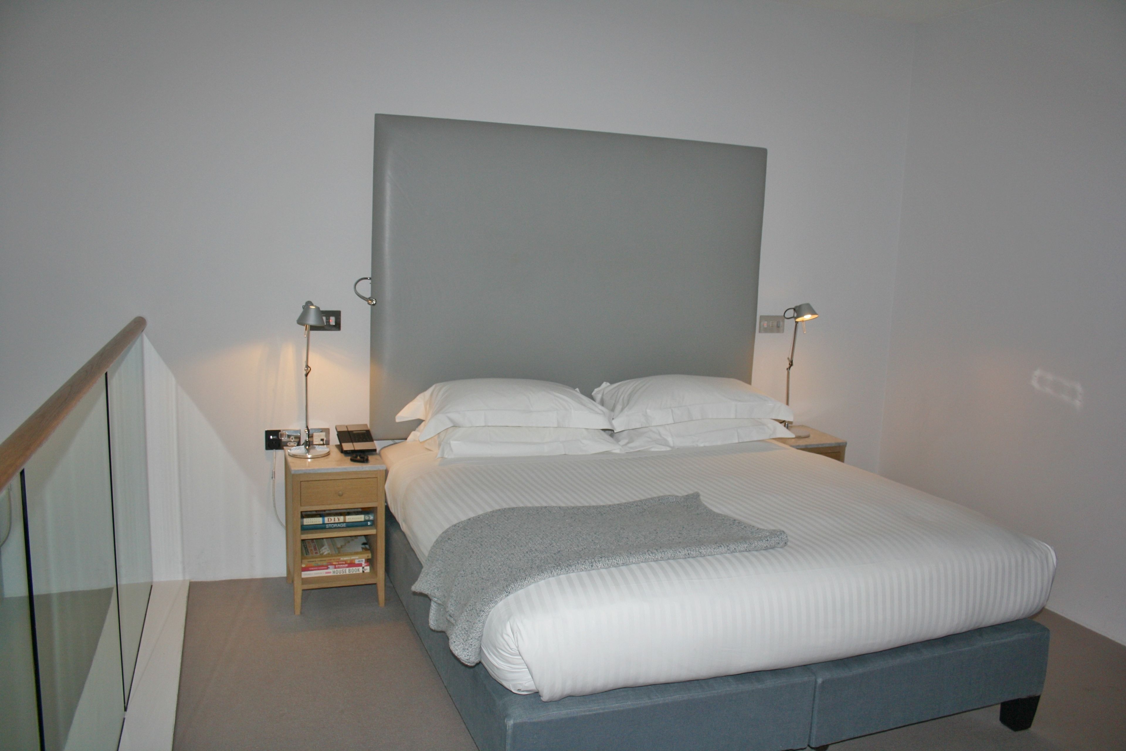 Ein ordentlich bezogenes Bett mit grauem Kopfteil, zwei Nachttischlampen und einem Nachttisch mit Lampe und Büchern.