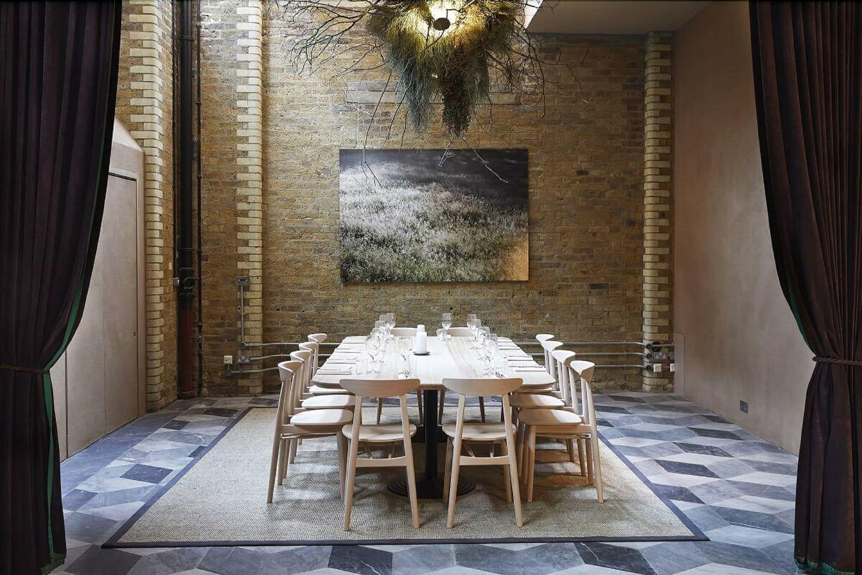 Uma sala de jantar com uma mesa comprida posta para refeição, rodeada por paredes de tijolo e um grande quadro abstrato.