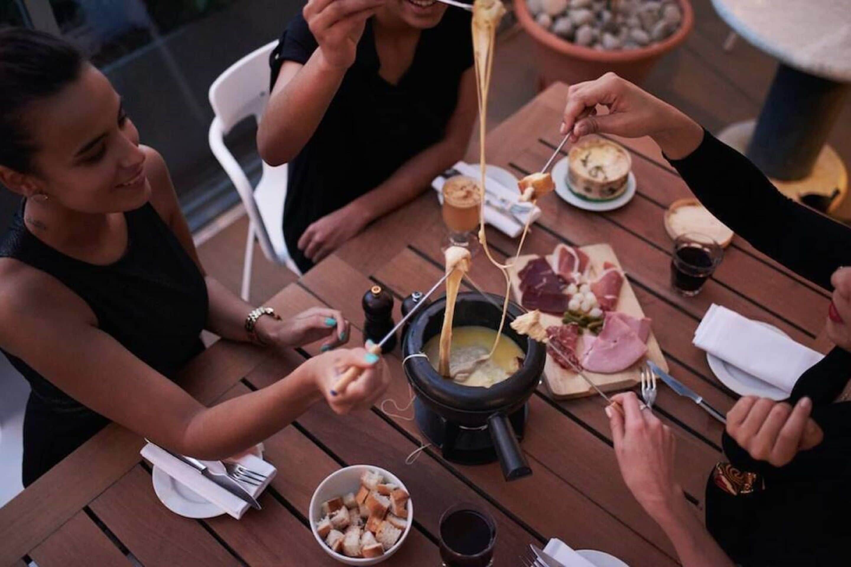A group of people enjoying a meal at an outdoor wooden table with a cheese fondue setup.