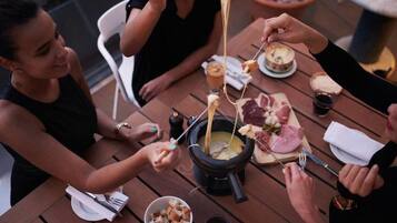 A group of people enjoying a meal at an outdoor wooden table with a cheese fondue setup.
