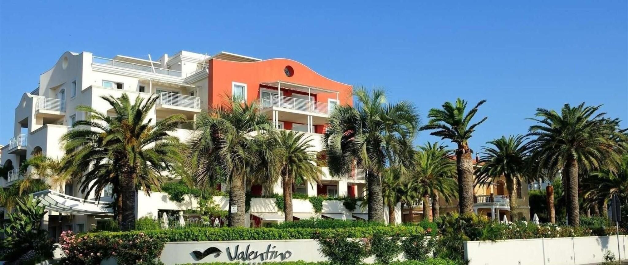 Ein Hotel mit der Aufschrift 'Valentino', umgeben von Palmen und blauem Himmel.