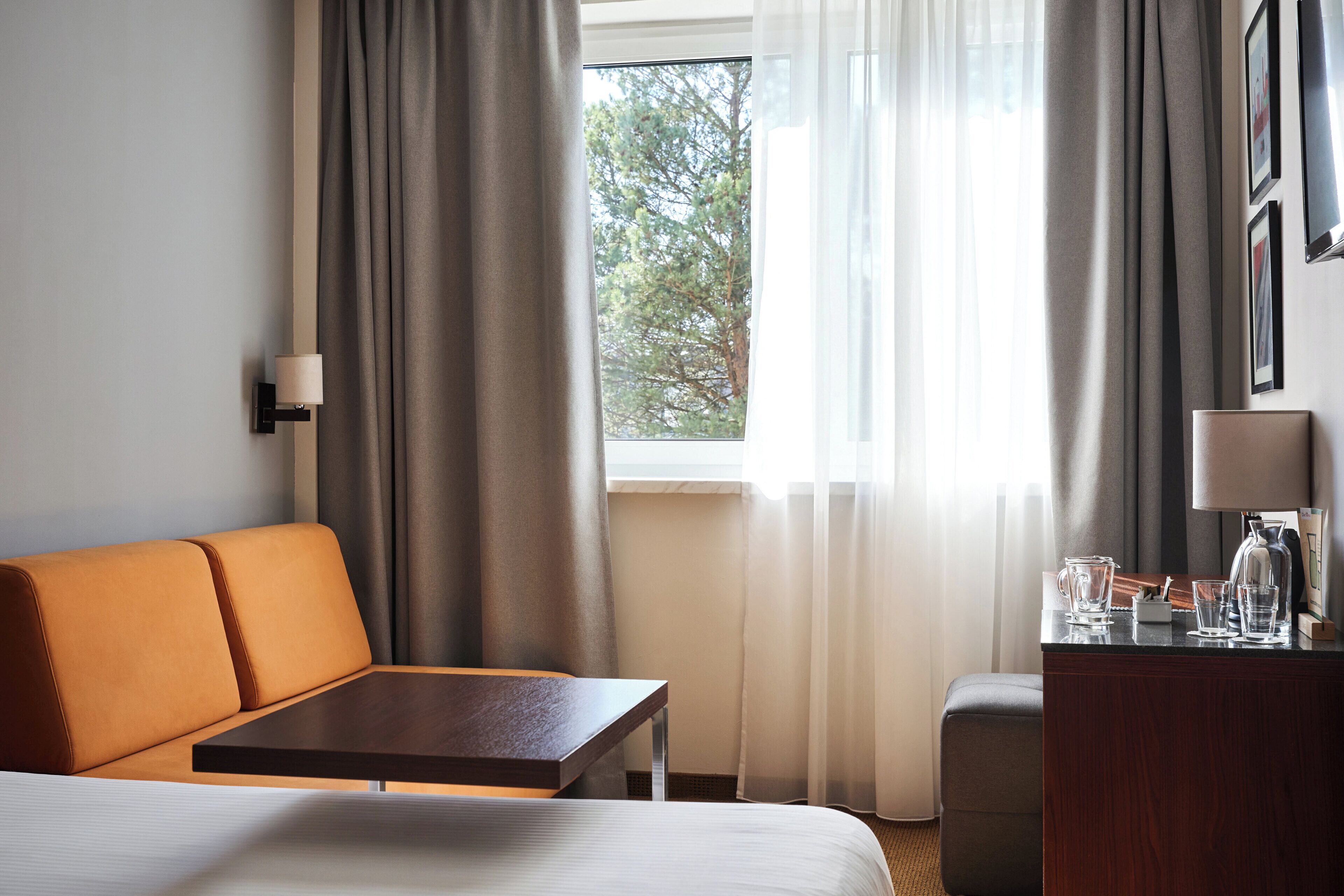 Una camera d'albergo con un letto, un divano, un tavolo, una scrivania, una lampada e una finestra con tende.