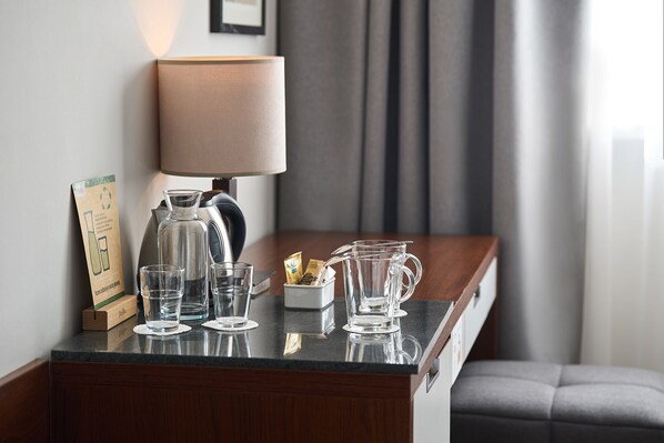 A hotel room with a coffee maker, glasses, a pitcher, and a sign on the counter.