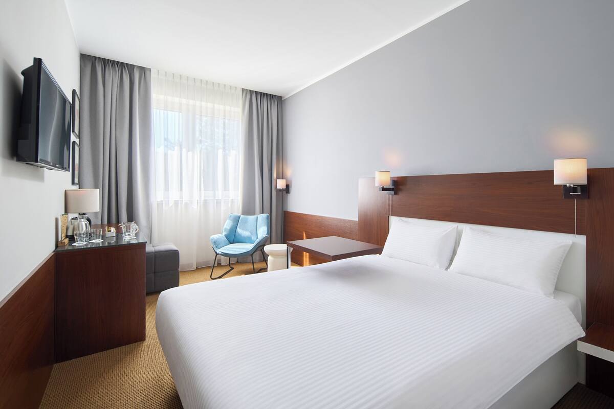 A hotel room with a large bed, a desk with a chair, a TV mounted on the wall, and a window with curtains.