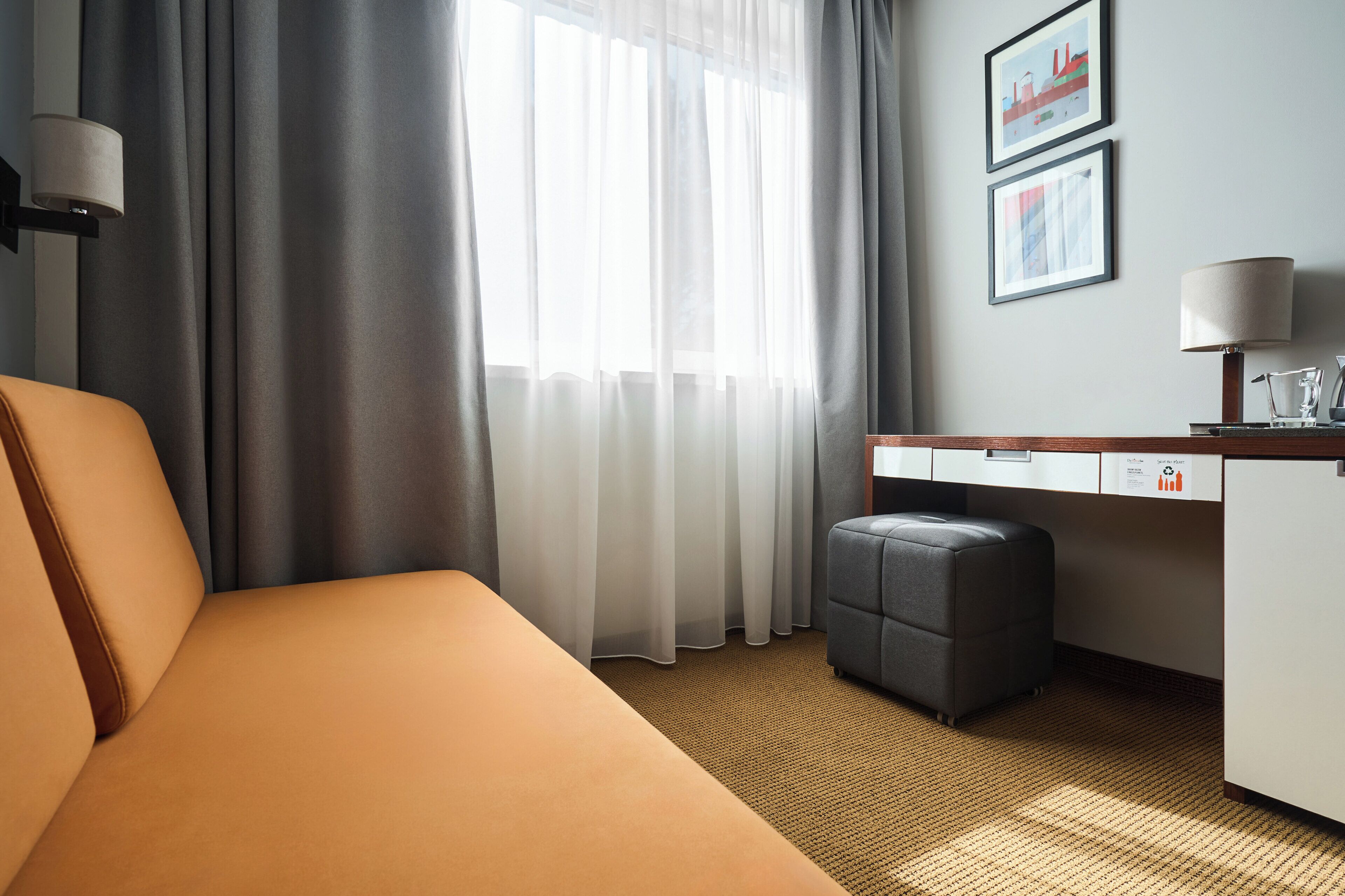 Una habitación de hotel con cama, escritorio, silla, lámpara y ventana con cortinas.