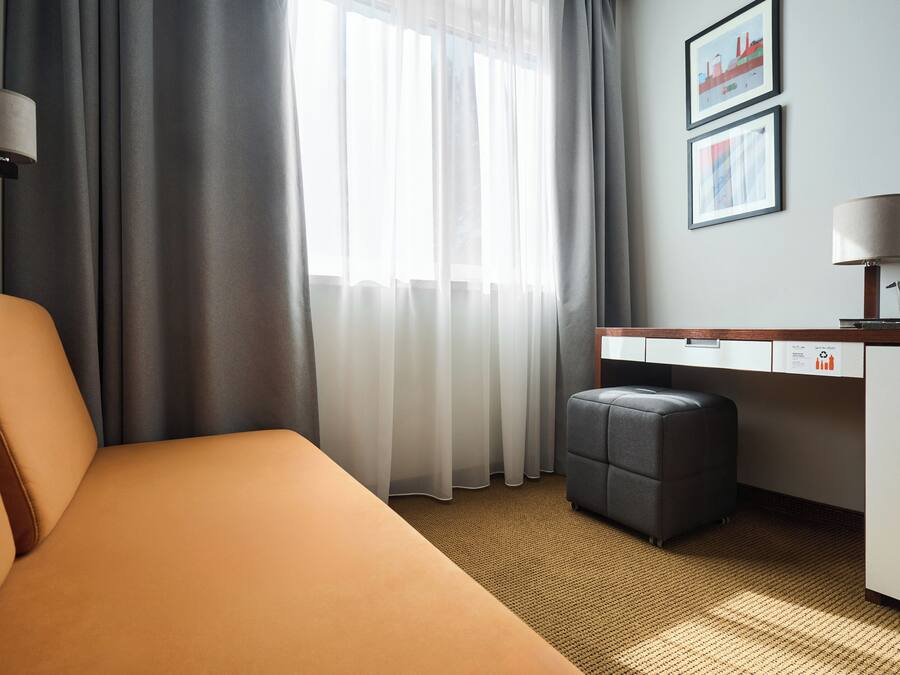 Una habitación de hotel con cama, escritorio, silla, lámpara y ventana con cortinas.