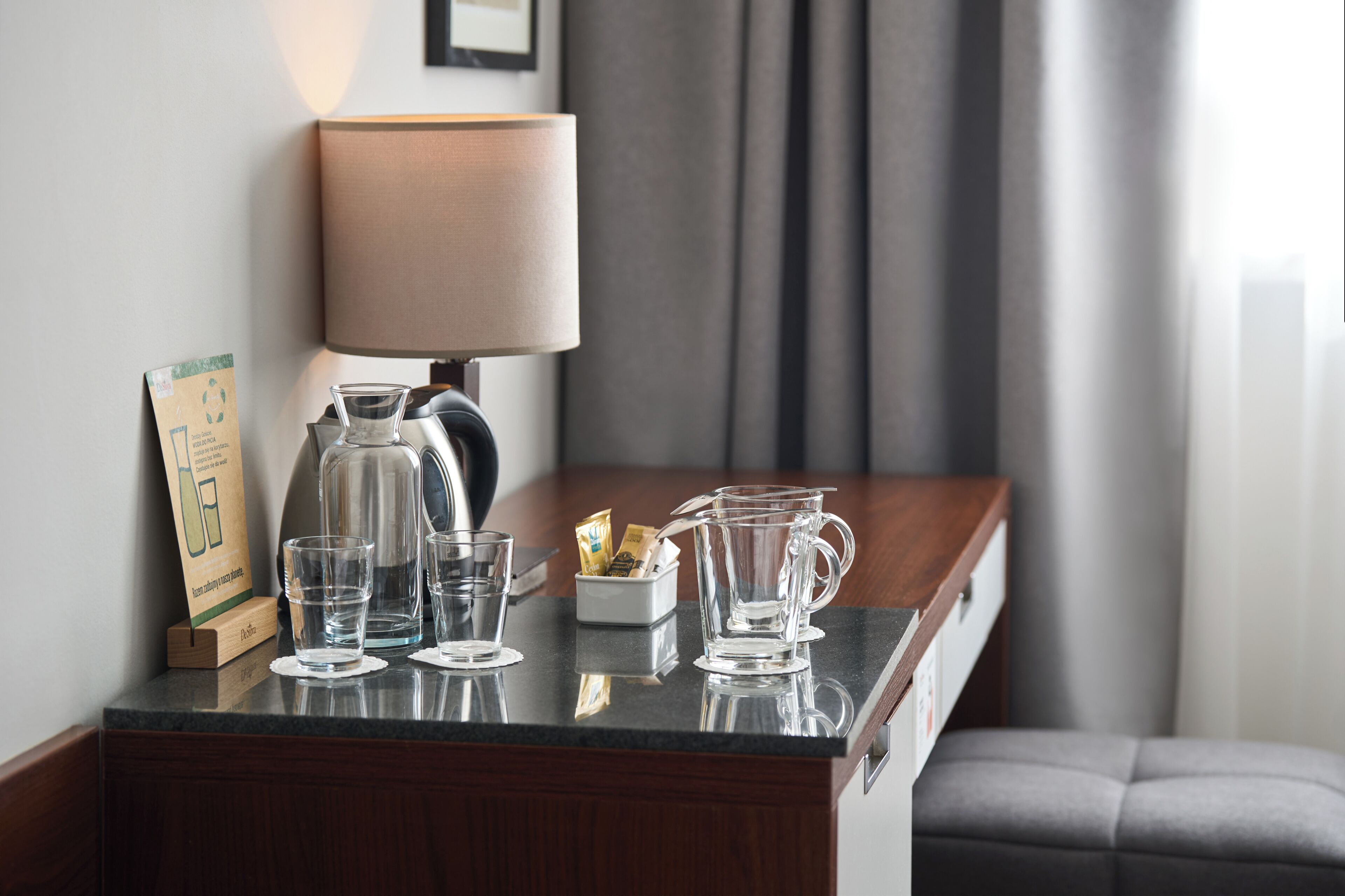 A hotel room with a desk, a lamp, a pitcher, glasses, and a sign.