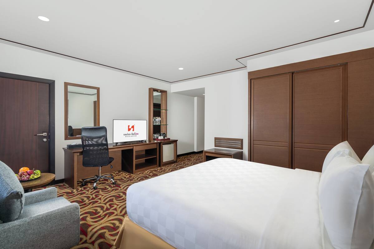 Une chambre d’hôtel avec un grand lit, un bureau avec un ordinateur, une chaise, une armoire et un miroir.