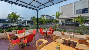 Un café al aire libre con sillas y mesas rojas y beige, rodeado de follaje y con vistas a un estacionamiento.