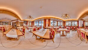 A spacious dining area with multiple tables and chairs, large windows offering a view of the outside, and a bar area visible in the background. - Loipenstub'n (Brixen im Thale)