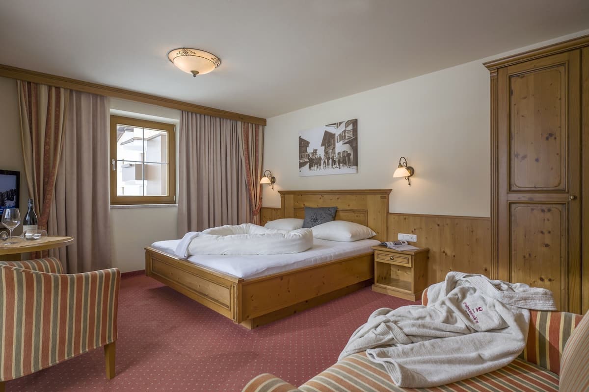 A hotel room with a wooden bed, a small bedside table, a chair, a wooden wardrobe, and a window with curtains.