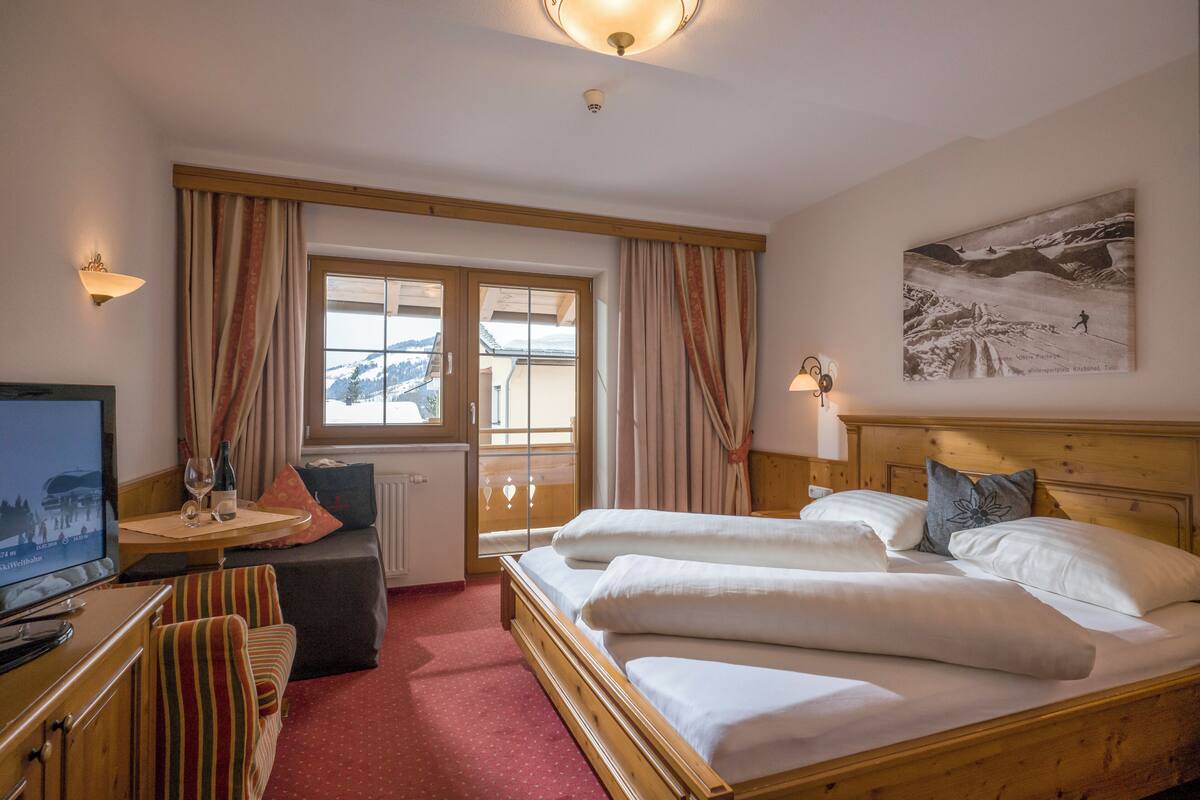 A hotel room with a bed, a TV, a small table with a bottle and glasses, a sofa, and a window with curtains.