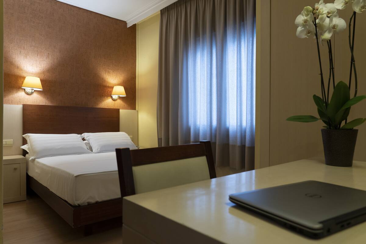 A hotel room with a bed, bedside lamps, a window with curtains, a table with a laptop, and a potted plant.
