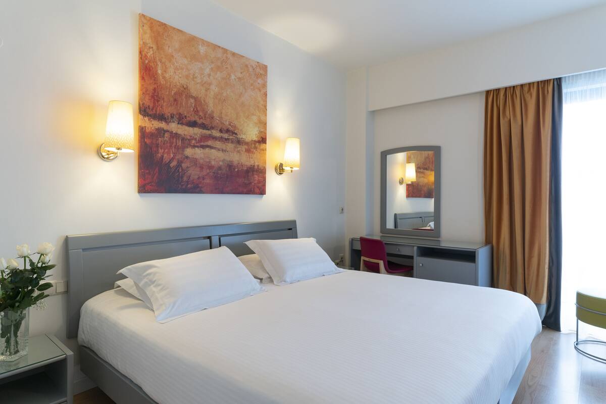 A hotel room with a large bed, bedside lamps, a desk, a chair, a vase with flowers, and a painting on the wall.