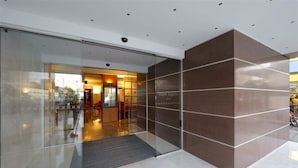Een moderne hotelentree met een glazen deur, een receptiegedeelte met zitplaatsen en een bruine wand met de naam van het hotel.