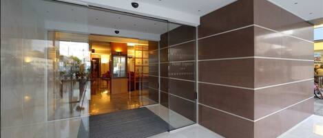 A modern hotel entrance with a glass door, a reception area with seating, and a brown wall with the hotel name.