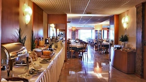 Een restaurantgedeelte in een hotel, met een buffetopstelling, een lange toonbank met eten en meerdere eettafels met stoelen.