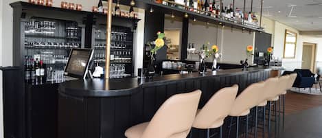 A bar area with a long counter, multiple shelves with bottles and glasses, and several cushioned bar stools.