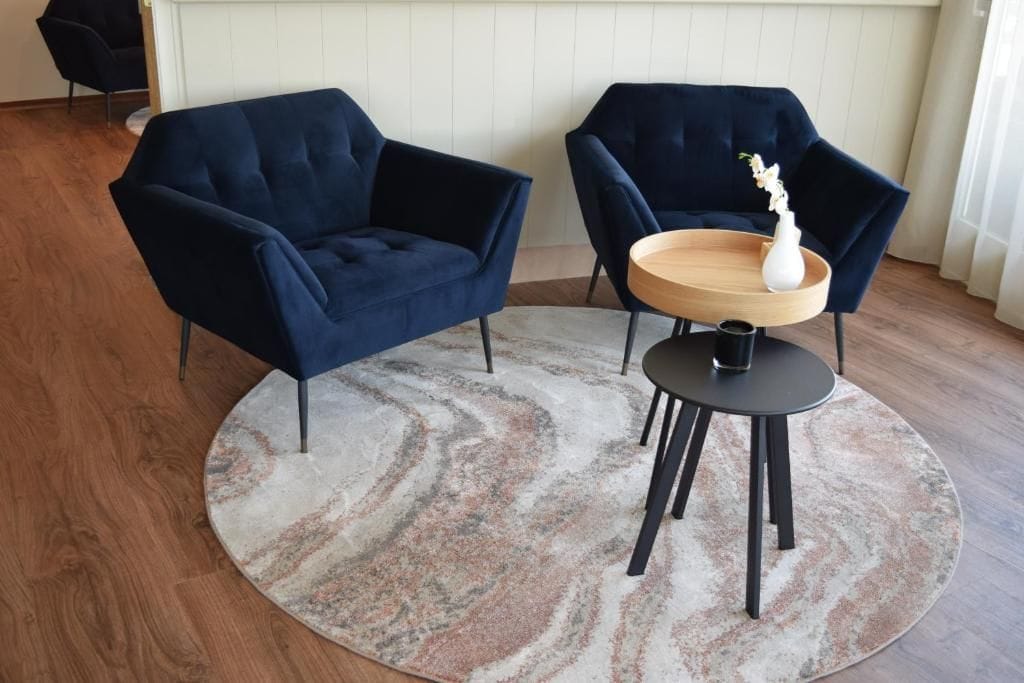 Una habitación con un sofá azul oscuro, una mesa redonda de madera con un jarrón blanco, y una alfombra estampada sobre piso de madera.