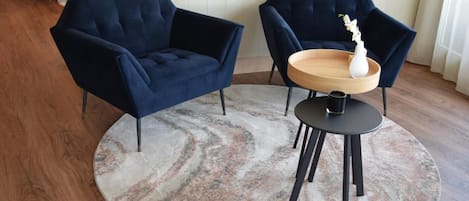 Um quarto com um sofá azul escuro, uma mesa redonda de madeira e um vaso branco, além de um tapete estampado sobre o piso de madeira.