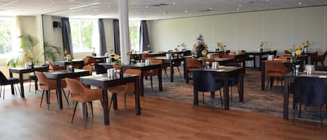 En rummelig spiseplads med flere borde og stole, store vinduer med gardiner og potteplanter.