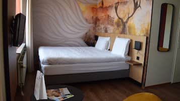Quarto de hotel com uma cama grande, uma mesa pequena, uma cadeira, televisão na parede e um quadro.