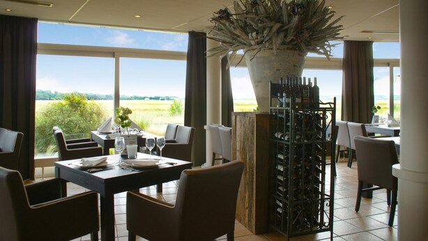 Een restaurant met een groot raamzicht op een veld, een wijnrek en een decoratieve plant.