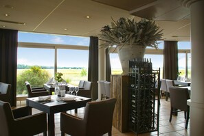 Un restaurant avec une grande fenêtre donnant sur un champ, un présentoir à bouteilles de vin et une plante décorative.