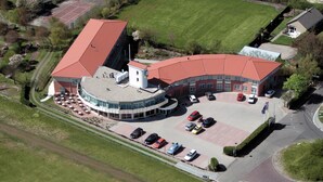 Luchtbeeld van een gebouw met een gebogen dak, een parkeerplaats en omliggend groen.
