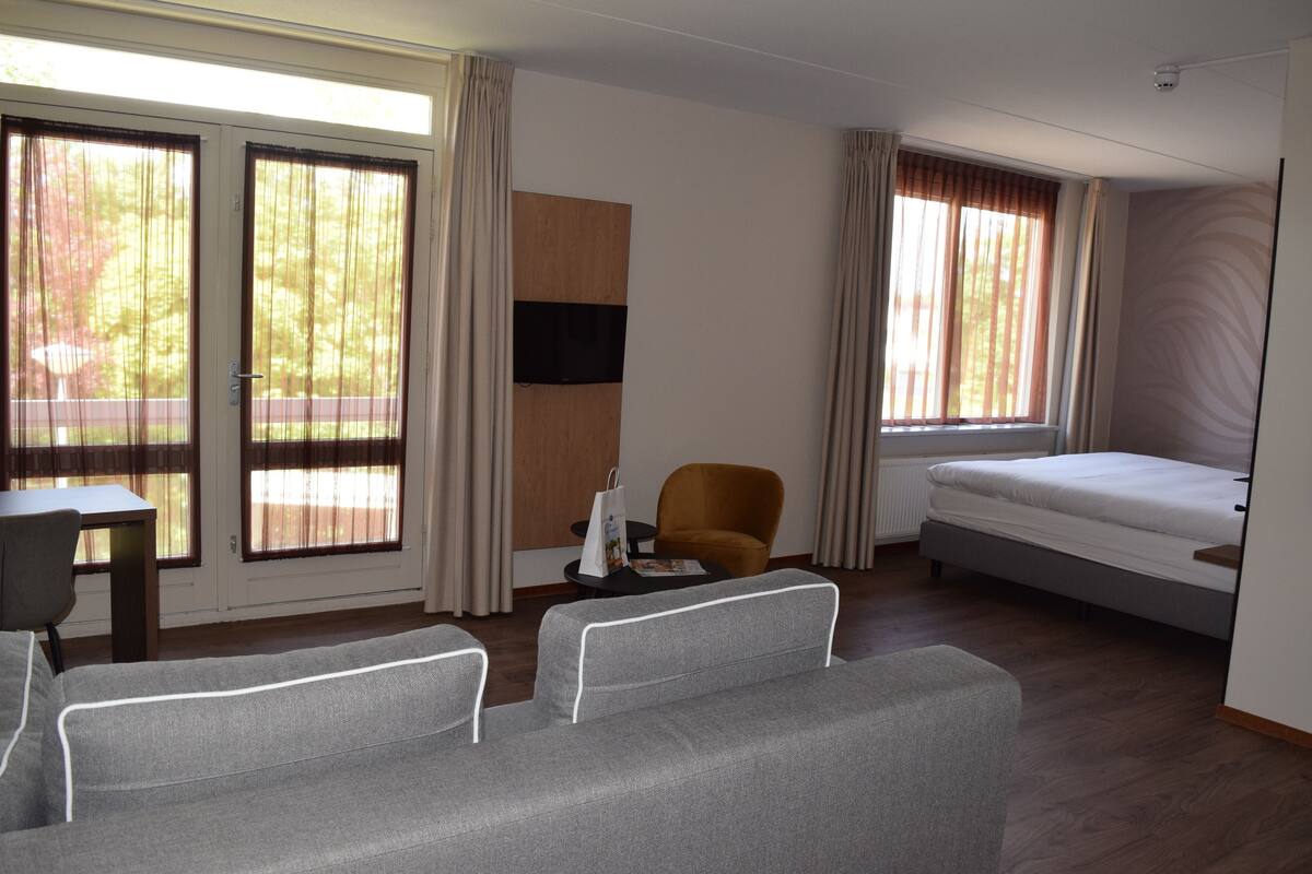 Een hotelkamer met een bank, een stoel, een bed, een televisie en grote ramen met gordijnen.