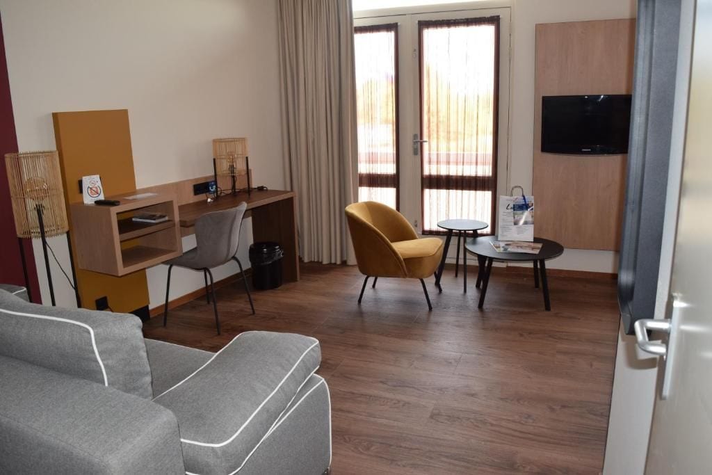 Une chambre d’hôtel comprenant un canapé, un bureau avec une chaise, une petite table avec une chaise, une télévision et une fenêtre avec des rideaux.