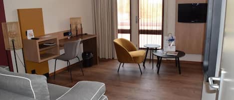 Une chambre d’hôtel comprenant un canapé, un bureau avec une chaise, une petite table avec une chaise, une télévision et une fenêtre avec des rideaux.