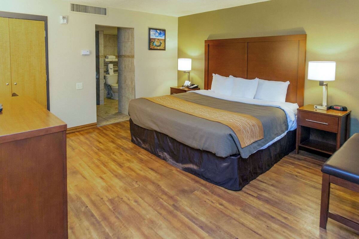 A hotel room with a wooden floor, a large bed with a headboard, two bedside tables with lamps, a wooden desk, and a door.