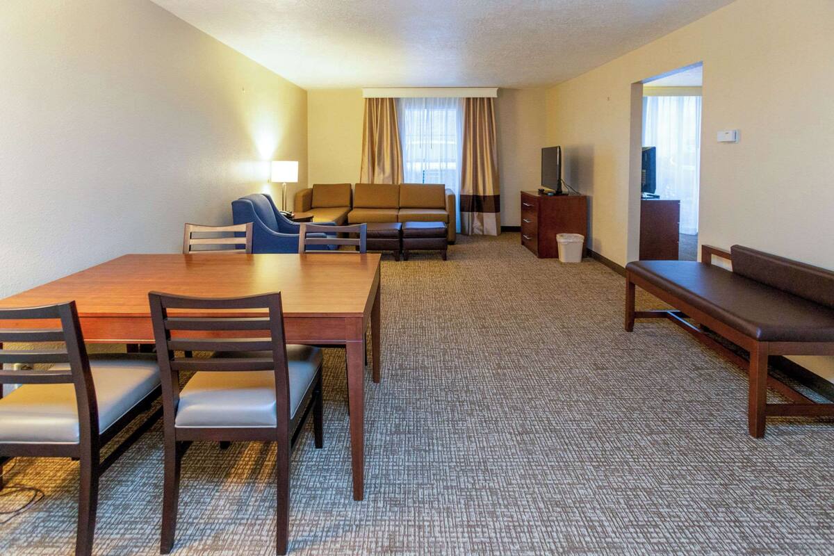A hotel room with a dining table, chairs, a sofa, a TV, and a trash bin.