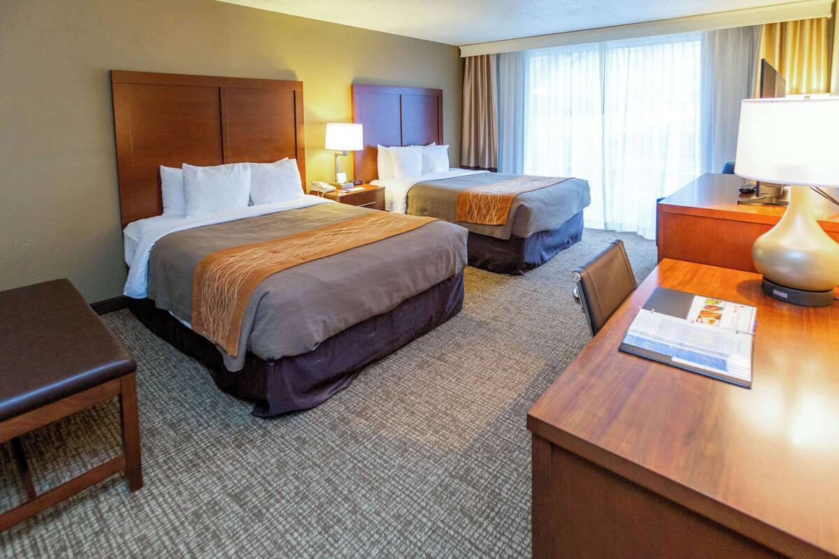A hotel room with two beds, a desk, a chair, and a lamp.