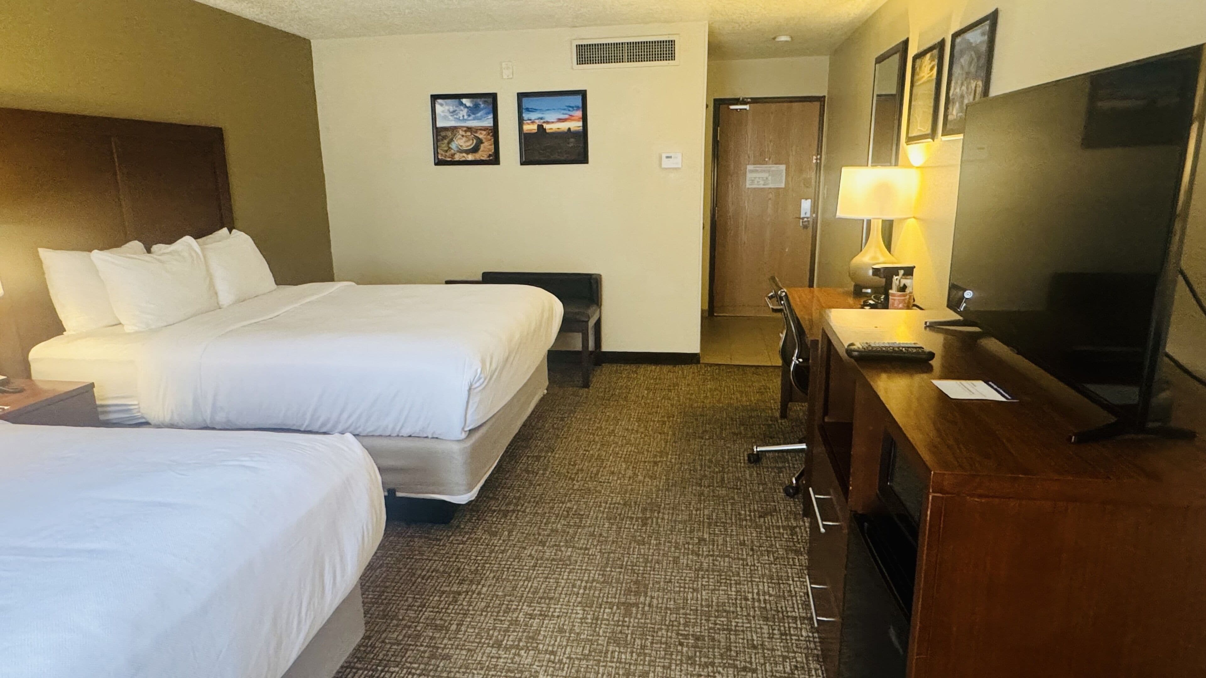 Een hotelkamer met twee bedden, een bureau met een lamp, een televisie en twee ingelijste foto’s aan de muur.