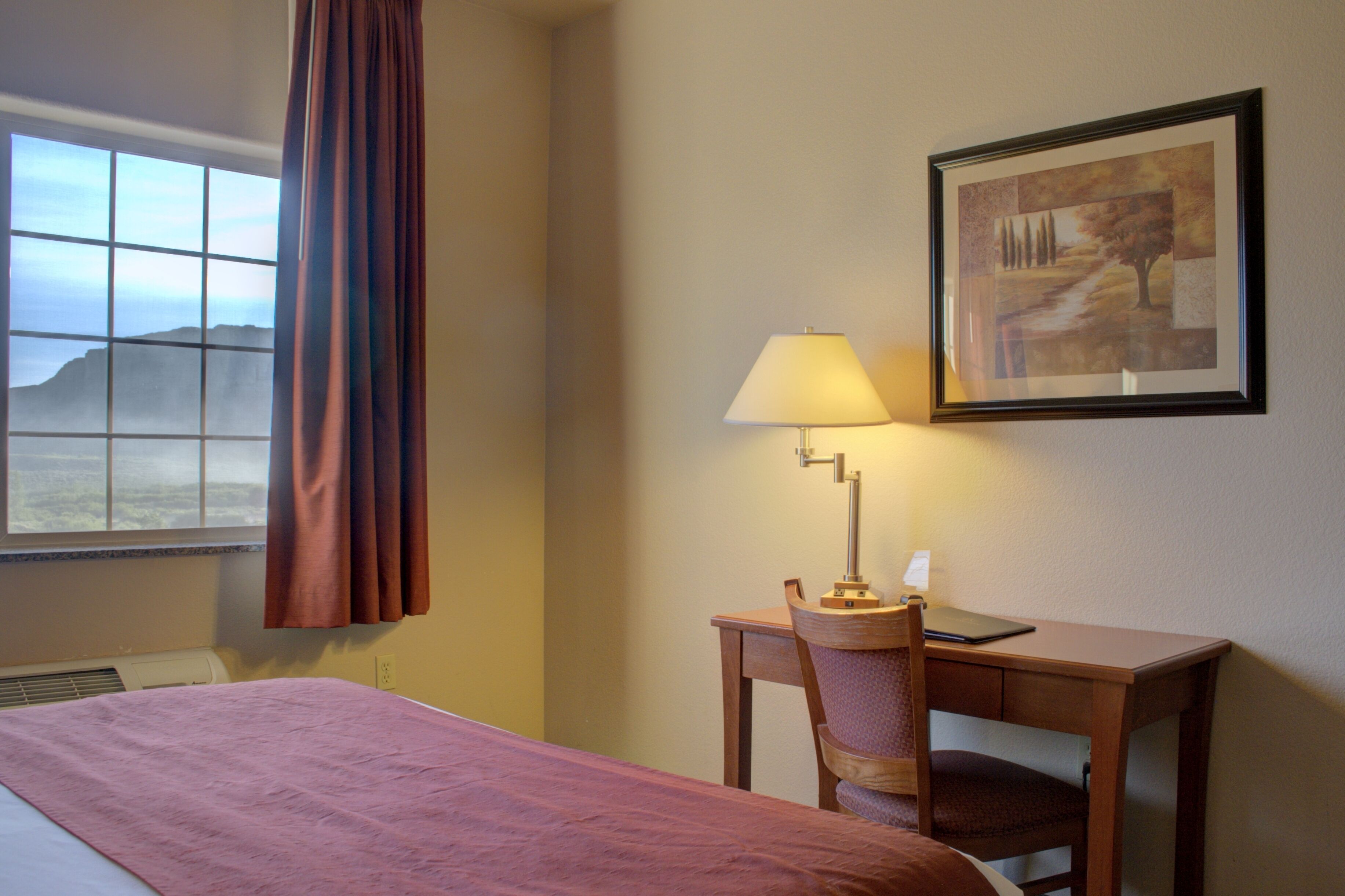 Een hotelkamer met een bed, een bureau met een lamp, een lijst met een foto aan de muur en een raam met gordijnen.