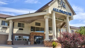A hotel with a prominent sign reading "Allington Suites." It features a covered entrance with columns and a brick base, large windows, and a clear blue sky in the background. - Allington Inn & Suites (Kremmling)