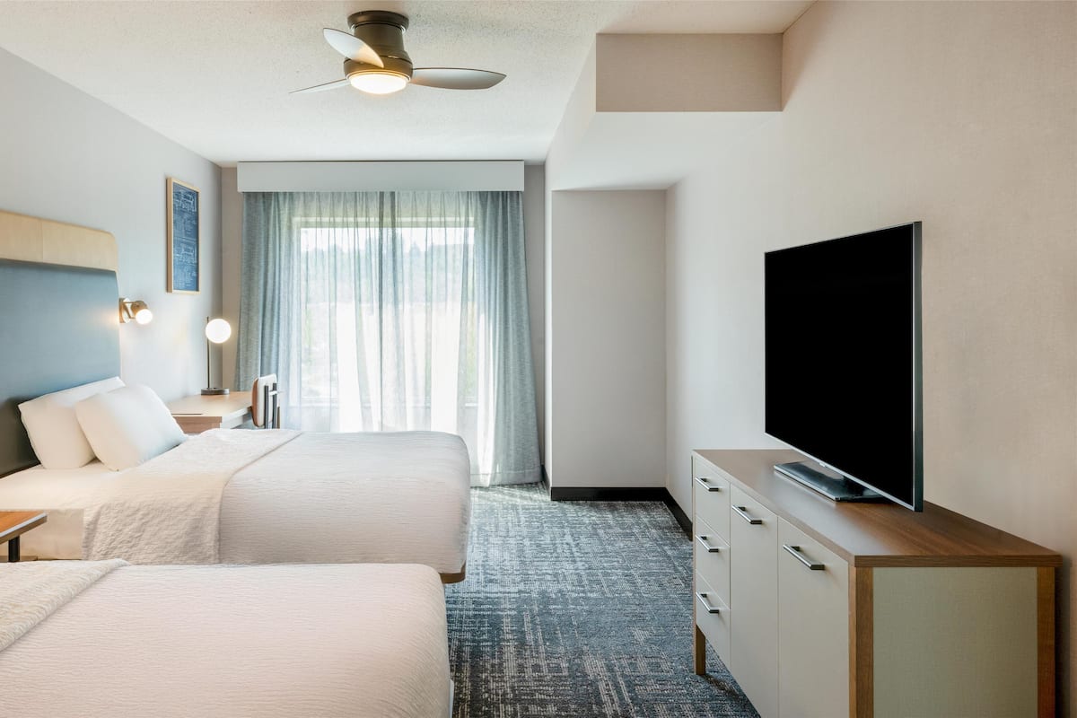 A hotel room with two beds, a flat-screen TV, a ceiling fan, and a large window with curtains.