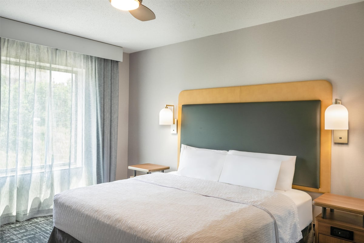 A hotel room with a large bed, two bedside tables, a headboard, and a window with curtains.