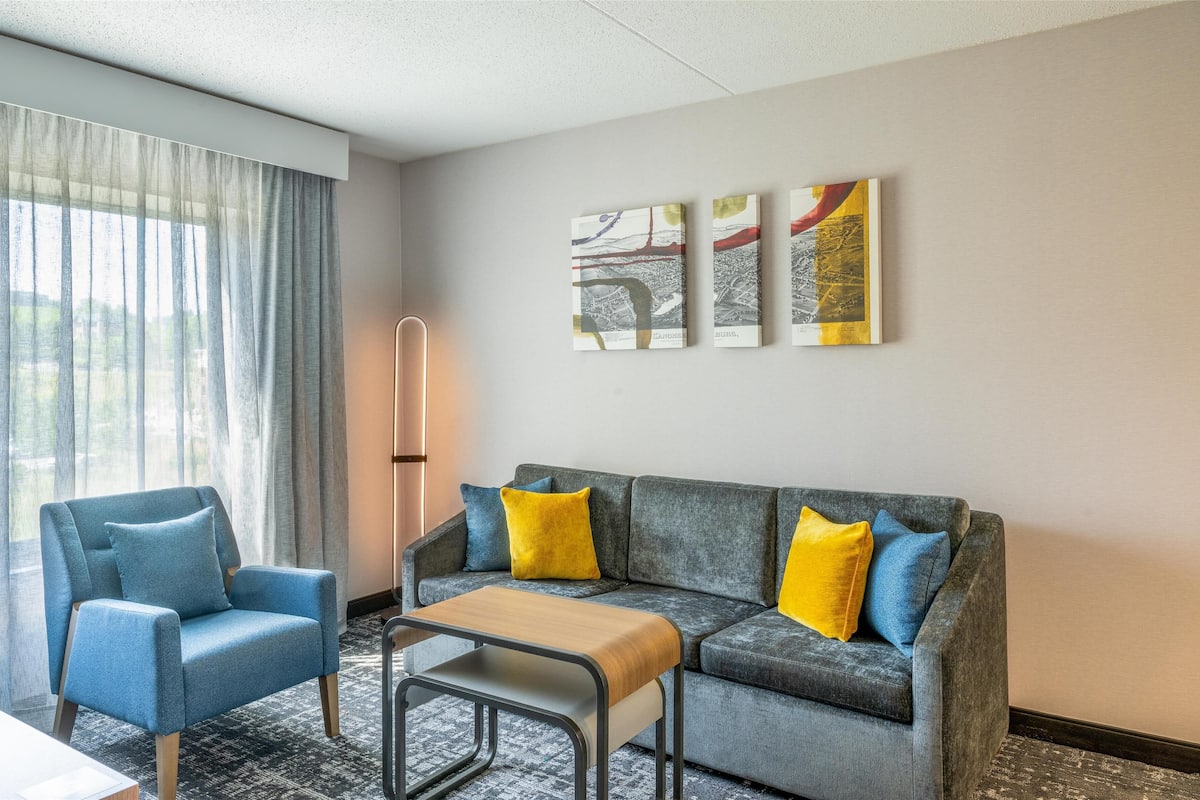 A hotel room with a grey sofa, a blue armchair, a wooden coffee table, and a floor lamp. There are abstract paintings on the wall.