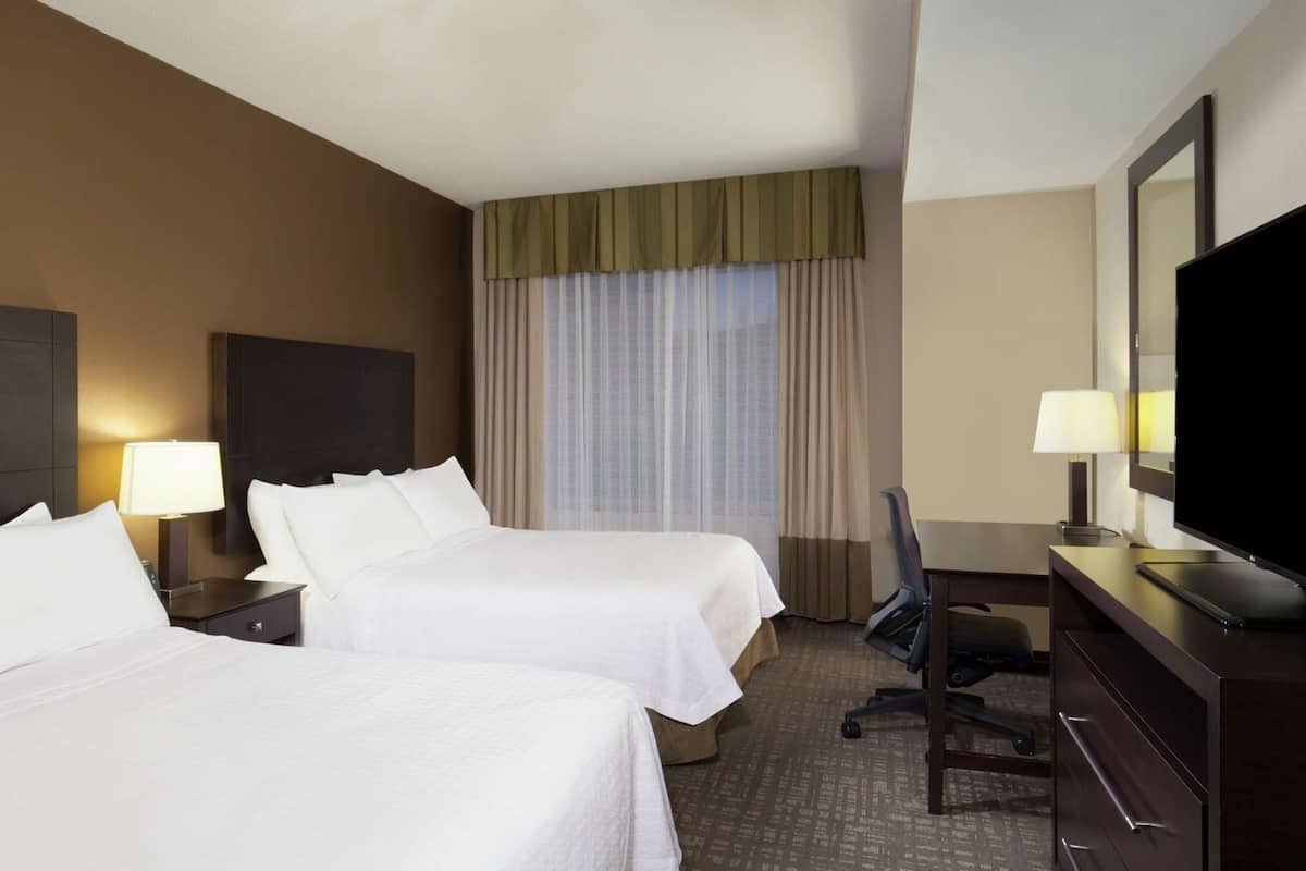 A hotel room with two beds, a desk, a chair, a TV, and a window with curtains.