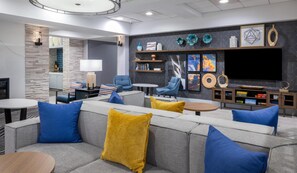 Een moderne woonkamer met een grijze bank, blauwe en gele kussens, een salontafel en een aan de muur bevestigde televisie.