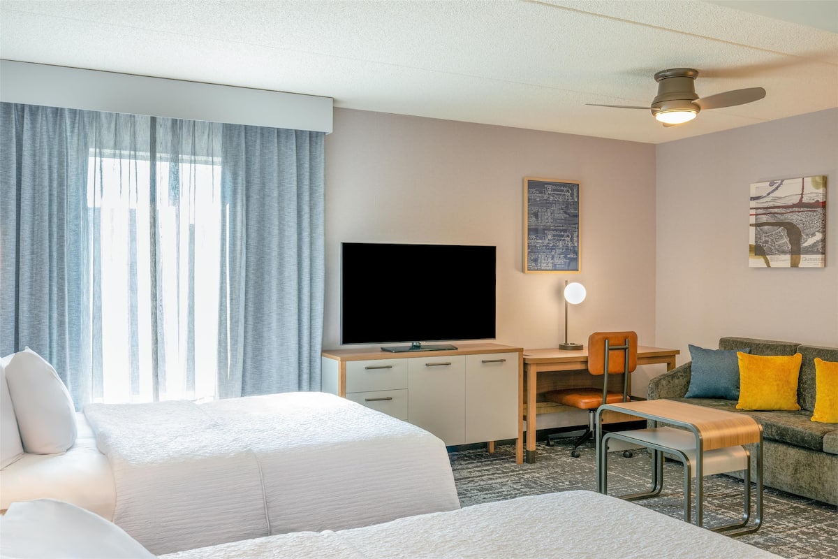 A hotel room with two beds, a desk with a TV, a chair, a sofa with cushions, and a ceiling fan.
