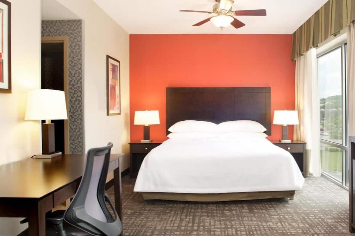 A hotel room with a bed, two bedside tables with lamps, a desk with a chair, a ceiling fan, and a large window.