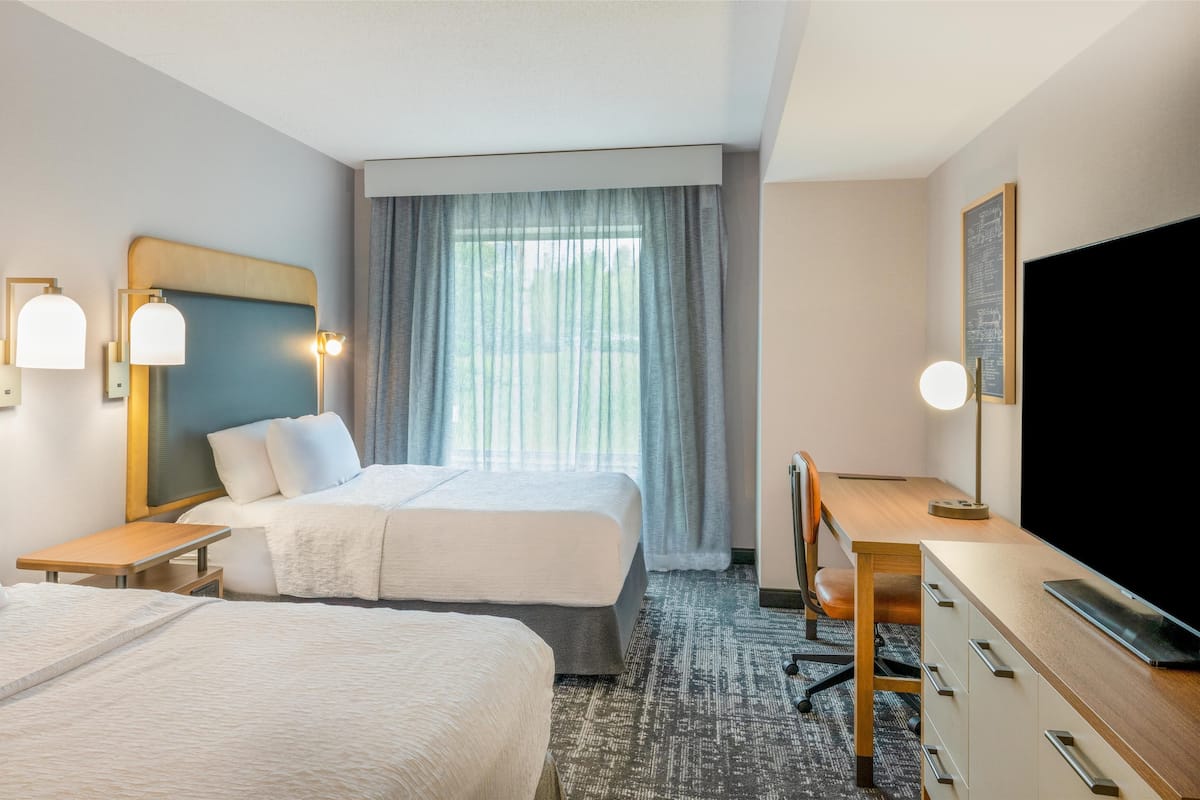 A hotel room with two beds, a desk, a chair, a TV, and a window with curtains.