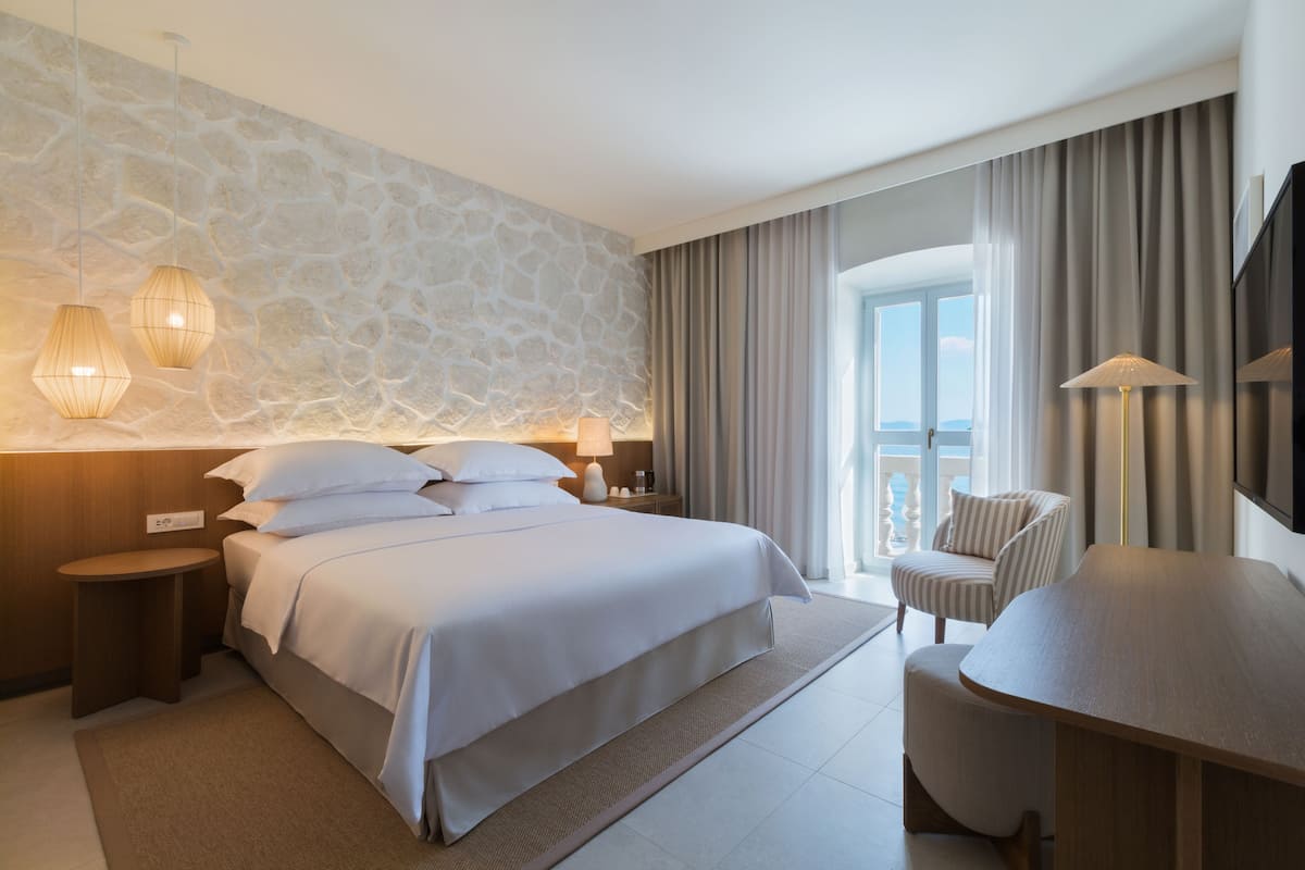 Et moderne hotelværelse med en stenvæg, en stor seng med hvide sengetøj, et siddeområde med et træbord og stole, samt en balkon med gardiner.